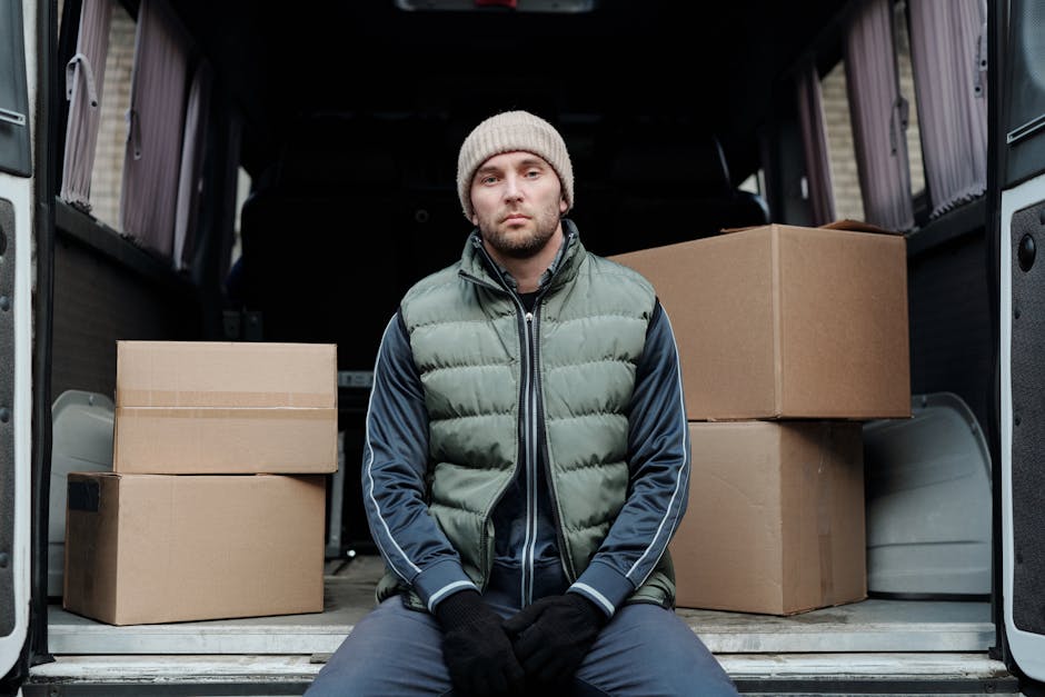 A young man wearing a grey knit beanie, a green puffer vest over a blue jacket, and black gloves, sitting inside the open rear of a large moving van. The van's interior is visible with several cardboard boxes of various sizes, some stacked and others placed on the floor, ready for or having recently been unloaded during a home relocation process. The van's side door is open, revealing a loading or unloading environment, with ambient natural light illuminating the scene. The boxes are wrapped in plain brown cardboard, and the moving van appears fully equipped for furniture transport and packing during a house removal, supporting professional moving services in East Dulwich, as offered by Man with Van East Dulwich.