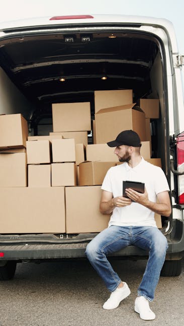 A man wearing a black cap, white t-shirt, and blue jeans sitting on the edge of an open van door during a home relocation process in daylight. The van's interior is filled with stacked cardboard boxes of various sizes, some taped and others open, containing household items and packaging materials. The boxes are arranged on the vehicle’s floor and on a small shelf, with a few larger boxes at the back. The man is holding a tablet device, possibly managing packing or loading details. The scene is set outdoors on a paved surface. This image reflects the loading process involved in furniture transport and packing during a house removal, with the van positioned close to a property for efficient moving logistics, captured by Man with Van East Dulwich, a provider in removals services.