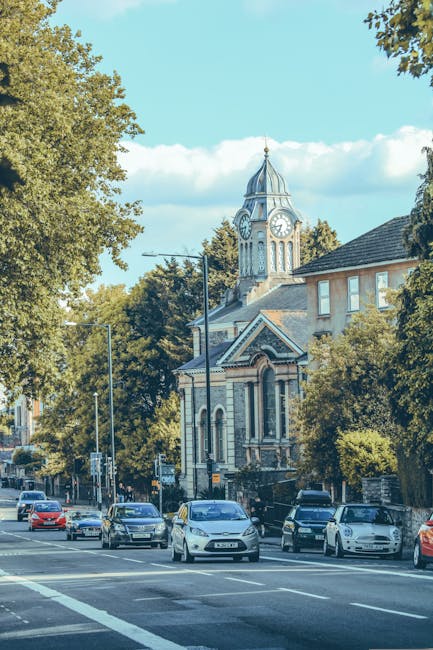 A view of a busy residential street in East Dulwich with parked cars lining both sides of the road, including sedans and hatchbacks in various colours, demonstrating an active home relocation environment. The road runs parallel to a sidewalk bordered by mature trees with lush green foliage, providing shade and a natural backdrop. In the background, a historic church with a tall clock tower and ornate architectural details rises above surrounding buildings, indicating a central community area. The church's clock shows the time as approximately 2:15. The sky is clear with some scattered clouds, and the scene captures daytime lighting with soft shadows cast by the trees and vehicles. The image highlights a typical move-in or move-out scene, where furniture, boxes, or packing materials may be transported in or out of nearby properties as part of a house removal service, with [COMPANY_NAME] specializing in local relocations in East Dulwich.
