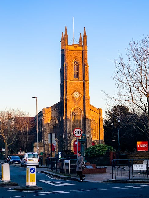 A tall, historic church building constructed from reddish-brown bricks, featuring a clock face on its front façade and narrow, arched stained glass windows. The church has a prominent square tower with pinnacles at the corners and a flagpole at the top. Surrounding the church are leafless trees, street signs including a 20 mph speed limit, and a few parked vehicles on the road. A person dressed in a coat is walking along the pavement, with some cardboard boxes and packing materials visible nearby, indicating a home relocation or moving activity. In the foreground, a curb with bollards and a fenced-off area mark the boundary of the street. This image supports content related to house removals, furniture transport, and moving logistics, as seen in the context of East Dulwich house moves when working with Man with Van East Dulwich.