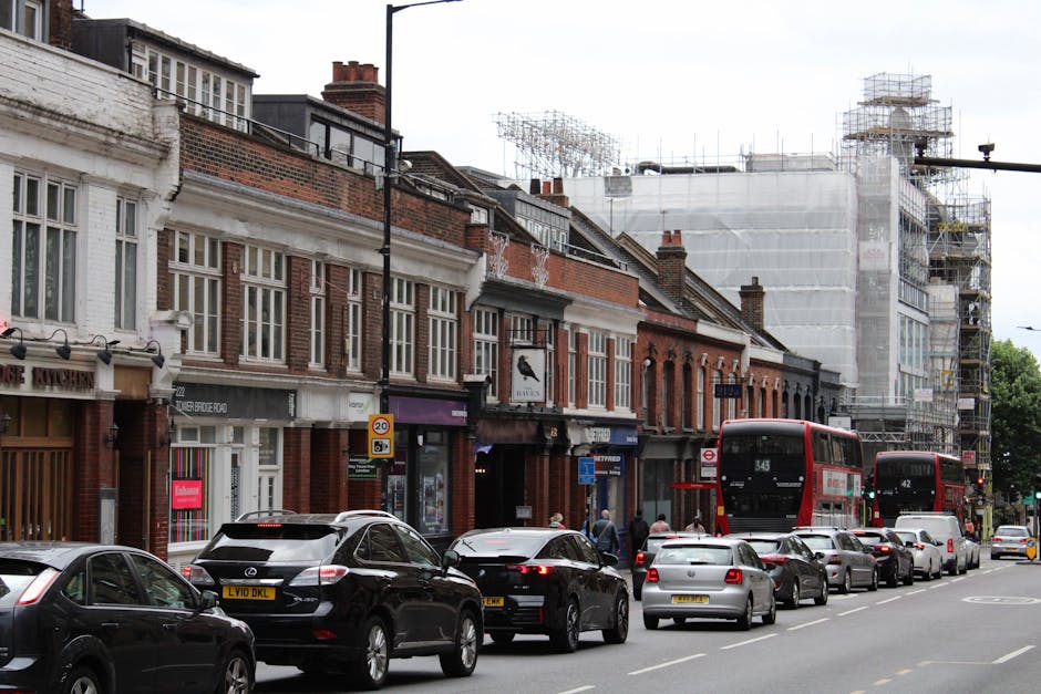 A view of a busy residential street in East Dulwich with parked cars lining both sides of the road, including sedans and hatchbacks in various colours, demonstrating an active home relocation environment. The road runs parallel to a sidewalk bordered by mature trees with lush green foliage, providing shade and a natural backdrop. In the background, a historic church with a tall clock tower and ornate architectural details rises above surrounding buildings, indicating a central community area. The church's clock shows the time as approximately 2:15. The sky is clear with some scattered clouds, and the scene captures daytime lighting with soft shadows cast by the trees and vehicles. The image highlights a typical move-in or move-out scene, where furniture, boxes, or packing materials may be transported in or out of nearby properties as part of a house removal service, with [COMPANY_NAME] specializing in local relocations in East Dulwich.