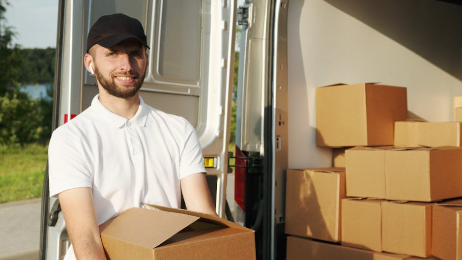A man in casual clothing is standing outside a white delivery van with an open rear door, loading and stacking cardboard boxes on the pavement. The boxes, made of brown cardboard, vary in size and are