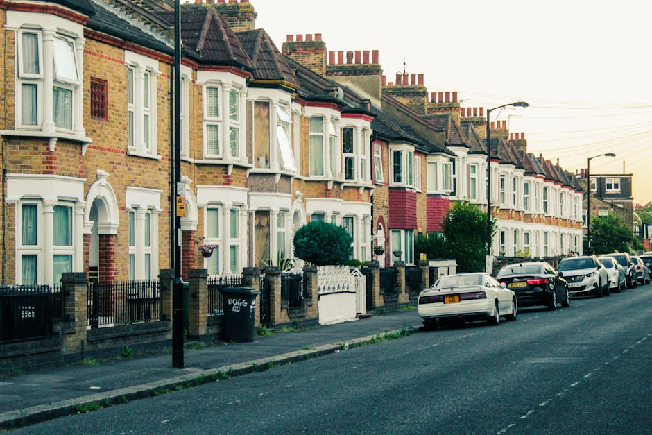 A row of brick terraced houses with multiple floors and large windows is visible along a paved sidewalk in an urban residential area. The buildings feature traditional Victorian-style architecture wit