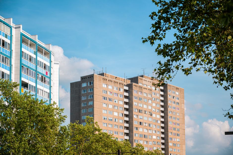 The image shows the exterior of two high-rise residential buildings in an urban area, with one building on the left featuring a modern glass and metal facade with blue accents, and the other on the right being a beige, concrete block structure with multiple rows of windows and small balconies. Both buildings are partially obscured by green leafy trees in the foreground. The sky above is bright blue with scattered white clouds, indicating a clear day. The scene captures the typical urban environment where home relocation and furniture transport might take place, possibly involving moving services such as those provided by Man with Van East Dulwich, who assist with packing and loading process within such residential complexes.