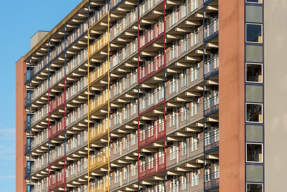 A close-up view of a modern multi-storey residential building exterior with a grid of balconies, each with metal railings painted in red, yellow, and dark blue. The balconies are arranged in vertical columns alongside large glass windows, reflecting the sky and surrounding environment. The building's façade includes a section with a peach-colored wall and a vertical row of windows on the right side. In the foreground, the image depicts the exterior of a property on North Cross Road, with a paved area and possibly some moving boxes or equipment nearby, suggesting an ongoing home relocation process. The scene is captured in natural daylight, highlighting the urban architecture typical of East Dulwich, suitable for visualising furniture transport, packing, and loading activities in the context of house removals, as handled by companies like Man with Van East Dulwich.