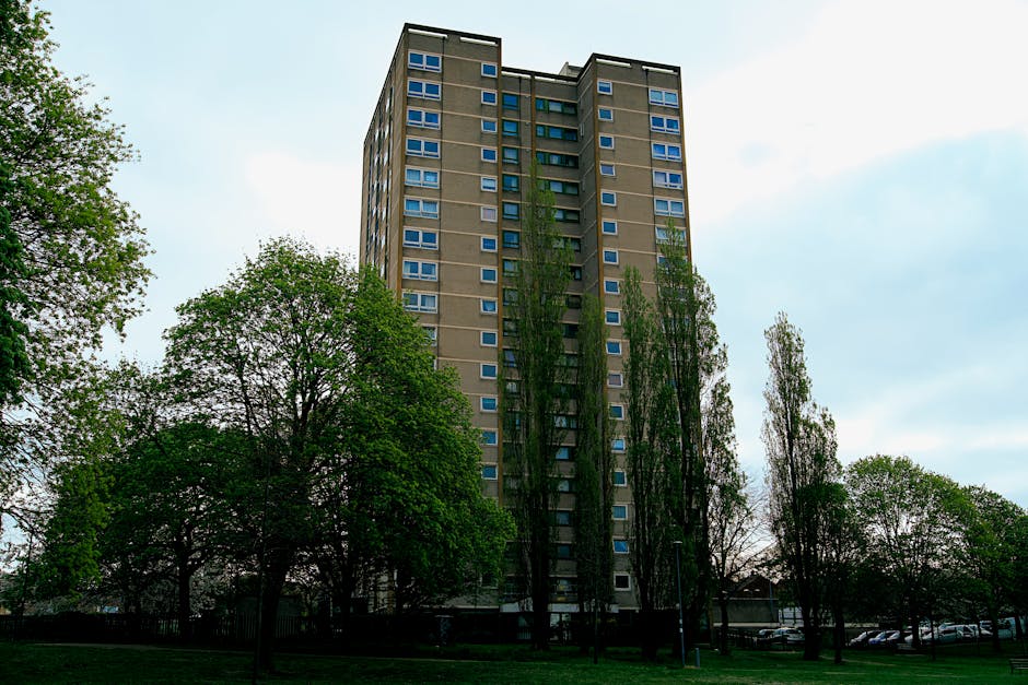 The image shows the exterior of two high-rise residential buildings in an urban area, with one building on the left featuring a modern glass and metal facade with blue accents, and the other on the right being a beige, concrete block structure with multiple rows of windows and small balconies. Both buildings are partially obscured by green leafy trees in the foreground. The sky above is bright blue with scattered white clouds, indicating a clear day. The scene captures the typical urban environment where home relocation and furniture transport might take place, possibly involving moving services such as those provided by Man with Van East Dulwich, who assist with packing and loading process within such residential complexes.